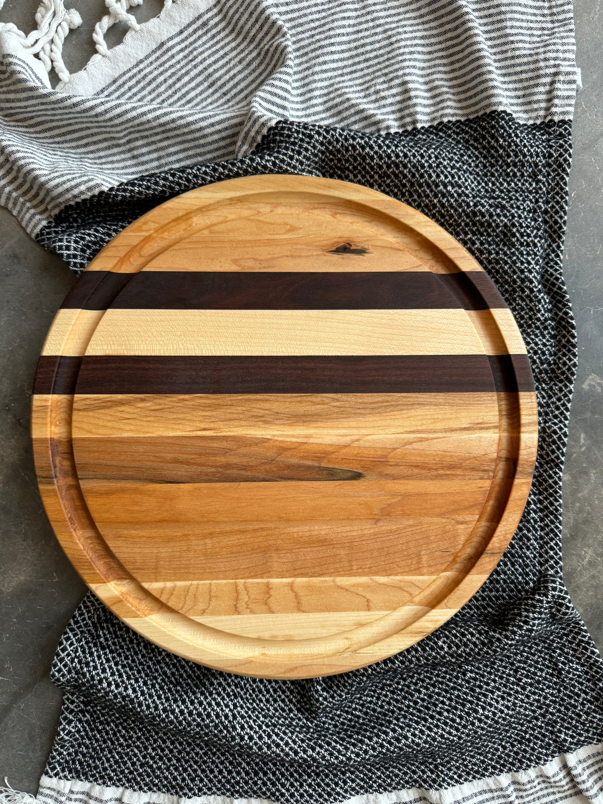 A round wooden cutting board with maple and walnut stripes. The board has a juice groove .