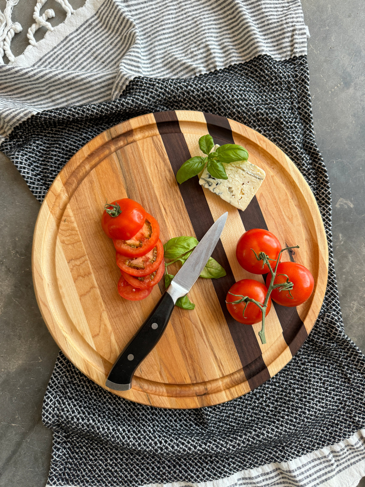 Round wooden edge grain cutting board with maple &amp; walnut stripes &amp; a juice groove. Topped with fresh tomatoes, basil and cheese, showcasing a stylish and functional serving or prep piece. Perfect for entertaining or gifting.