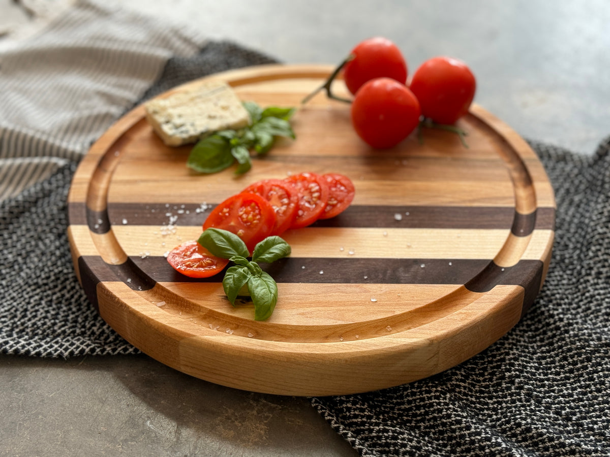 Round wooden edge grain cutting board with maple &amp; walnut stripes, personalized engraving, &amp; a juice groove. Pictured from a side view to appreciate the board thickness and quality. A stylish &amp; functional serving or prep piece.