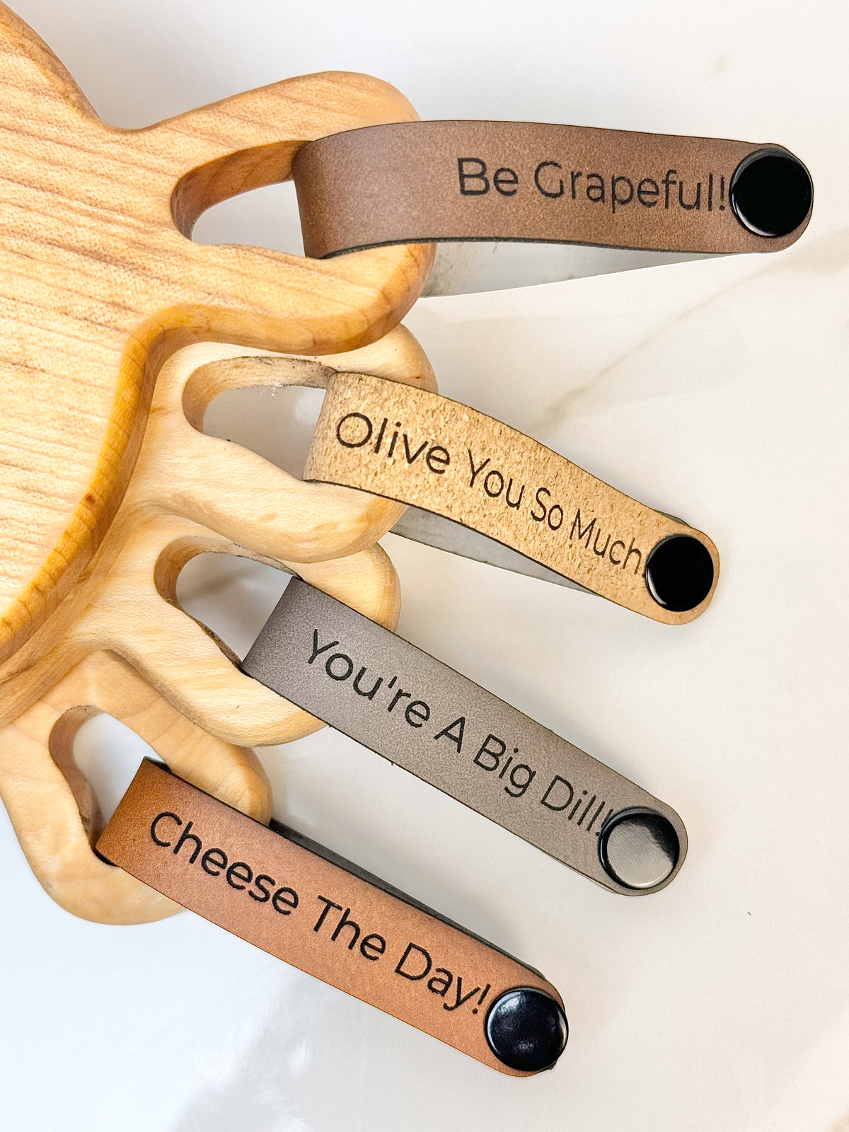 Maple wooden coaster single serve mini charcuterie board set with vegan leather straps engraved with puns: Be Grapeful!!, Olive You So Much, You're A Big Dill!, and Cheese The Day! Coasters have a natural finish.