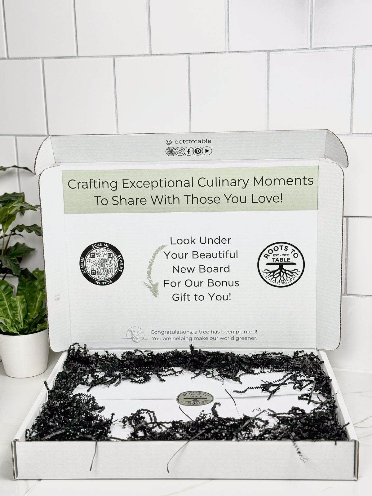 This image showcases how the charcuterie and cutting boards are packaged and presented as a gift It is wrapped in white paper held together with a logo sticker. Gaps are filled with black crinkle paper. Beautiful gift presentation
