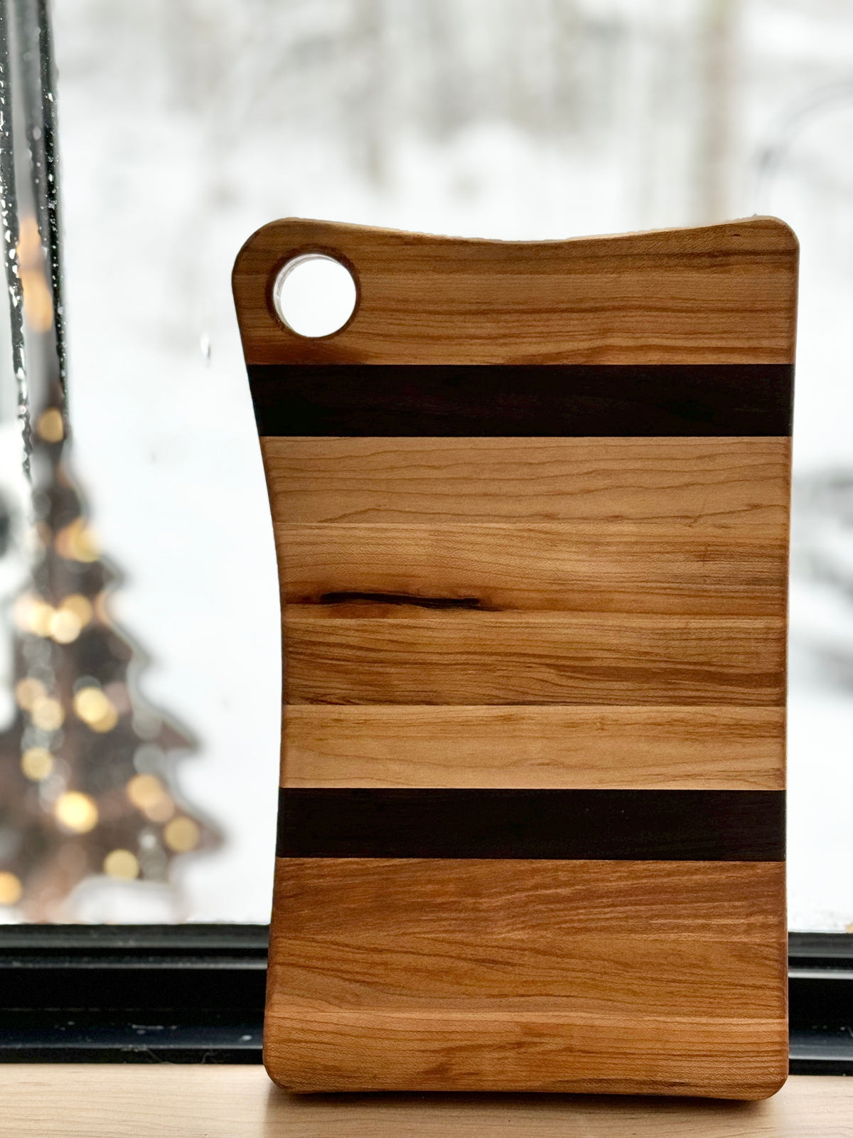 A 12-inch by 7-inch wooden cutting board with alternating maple and walnut wood stripes, has a built-in hole for easy storage. Customization available
