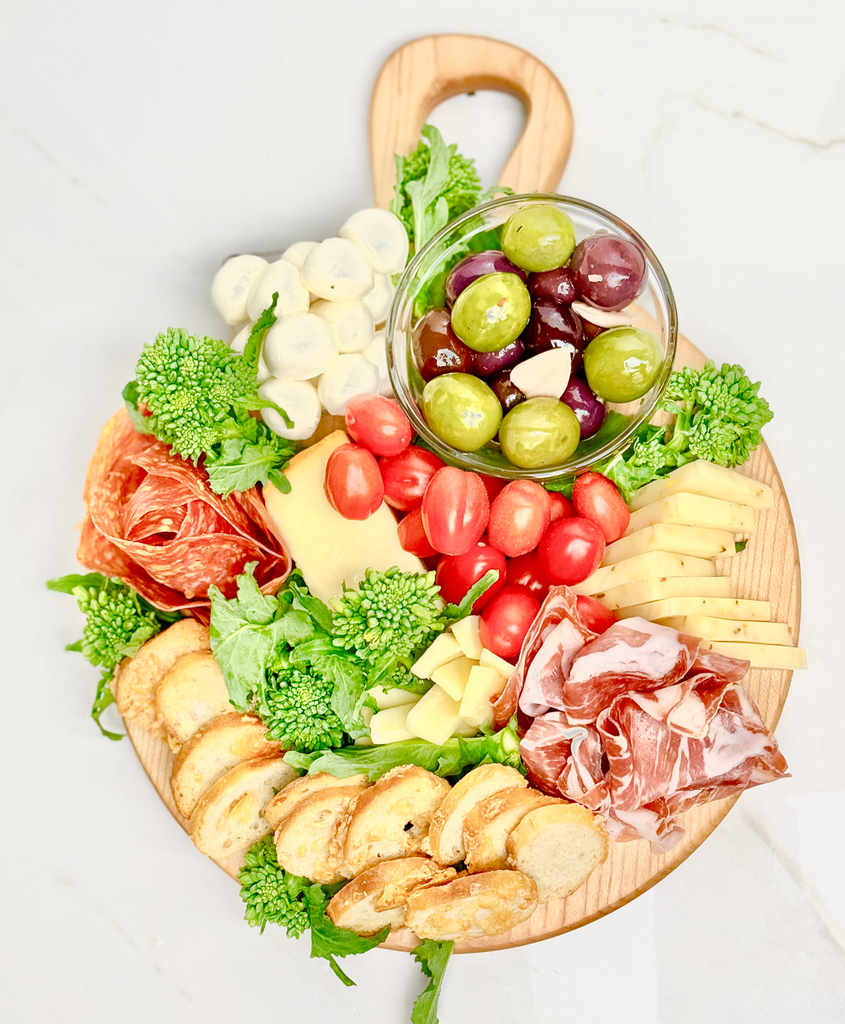 A round maple charcuterie board with handle is displayed loaded with deletable snacks.