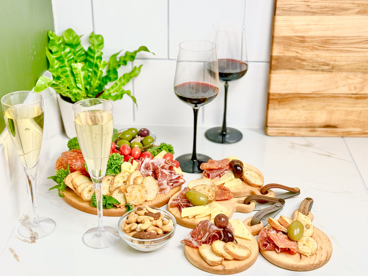 A stylish charcuterie spread on round wooden maple boards with meats, cheeses, olives, crackers, and nuts, accompanied by two champagne flutes and two glasses of red wine. A maple cutting board and a green plant decorate the bright marble countertop setting. This image showcases a medium sized round board that comes with 4 smaller individual round boards with witty food puns printed on attached vegan leather tags.