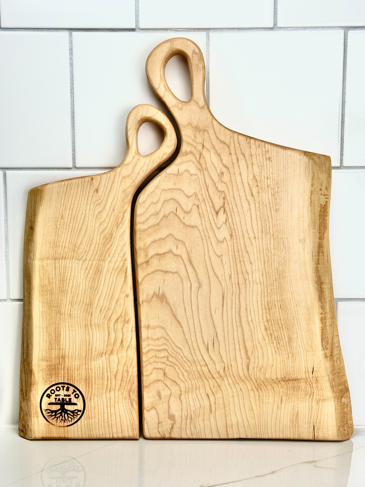 A pair of handcrafted wooden live edge boards engraved with Charlene & Jason, designed to nestle together. The handles fit snugly, symbolizing the intimate connection of a woman resting her head on a man's shoulder, creating one harmonious piece.
