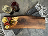 A handcrafted, solid walnut live edge  charcuterie board featuring rich natural wood grain, smooth edges, resting on a textured concrete surface, two glasses of wine and some food items such as olives a cheese rose, two meat roses crackers and rosemary garnish/