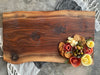 A live-edge walnut face grain slab charcuterie board featuring a natural grain pattern with natural imperfections and character. Topped with crackers, olives, salami roses, and cheese, it rests on a striped and black textured cloth on a rustic concrete surface.