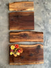 A display of four live-edge wooden charcuterie boards in varying sizes and shapes, showcasing rich walnut face grain patterns with natural imperfections. The bottom board is styled with an elegant charcuterie arrangement featuring salami, cheese, crackers, olives, and garnishes.