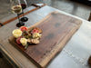 A live-edge walnut charcuterie board, a natural face grain slab, featuring personalized engraving, elegantly arranged with cheese, crackers, olives, and meats. Two glasses of wine sit nearby on a vintage metal table, creating a sophisticated dining atmosphere.