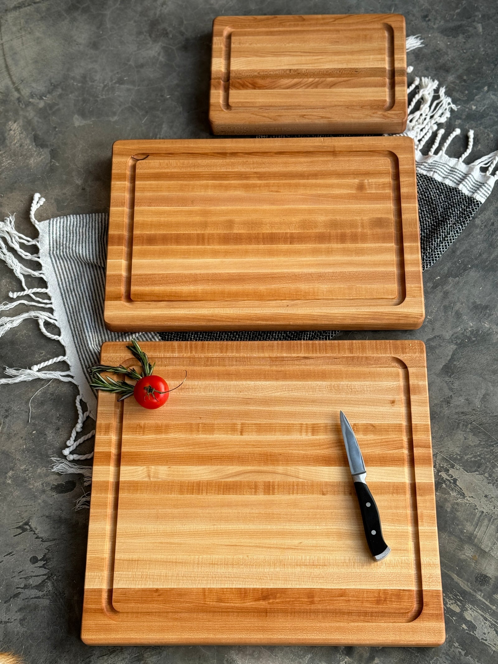 A set of three handcrafted maple cutting boards in varying sizes with juice grooves, displayed on a textured surface with a striped kitchen towel. A small knife and a fresh tomato with rosemary garnish sit on the largest board, highlighting its functionality and elegance.