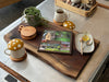 A large natural live edge face grain walnut charcuterie board showcased as a decorative item, it sits on a silver metal coffee table. Upon the board there is a string of pearls plant, two wooden coasters that look like mini charcuterie boards, a book, and a tray with honey and a spoon for the two cups of tea severed in mushroom tea cups.