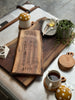 A cozy tabletop setup featuring an engraved live-edge wooden cribbage board atop a larger walnut charcuterie board. Surrounding the boards are two mugs of tea with mushroom top steeping lids, a honey jar with a dipper, and a potted plant.