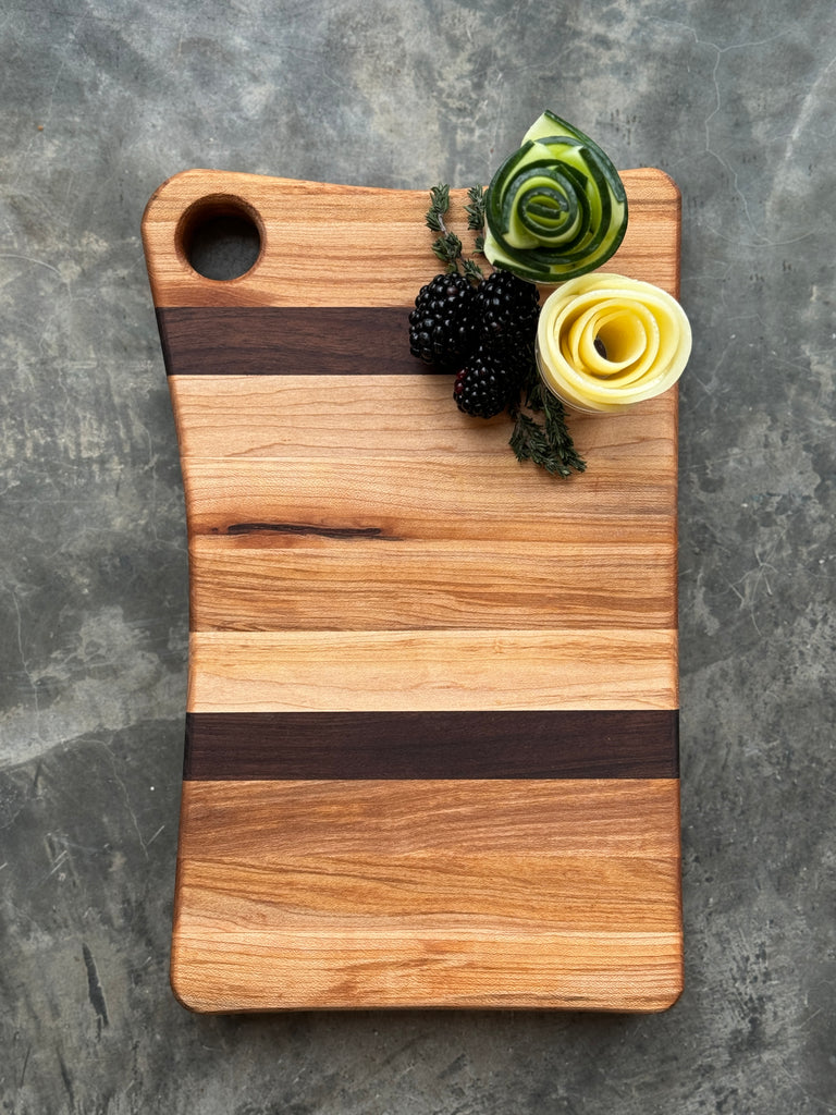 A handcrafted cutting board made of maple and walnut with a striped pattern, featuring a round handle cutout. Topped with decorative cucumber and cheese roses, blackberries, and herbs, placed on a textured cloth background.