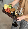 A women holds a dark walnut appetizer tray featuring a wine glass holder, crackers, blackberries, salami, cheese, and garnished with fresh herbs, showcasing an elegant portable charcuterie experience.