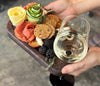 Close-up of hands holding a dark walnut appetizer tray featuring a wine glass holder, crackers, blackberries, salami, cheese, and garnished with fresh herbs, showcasing an elegant portable charcuterie experience.