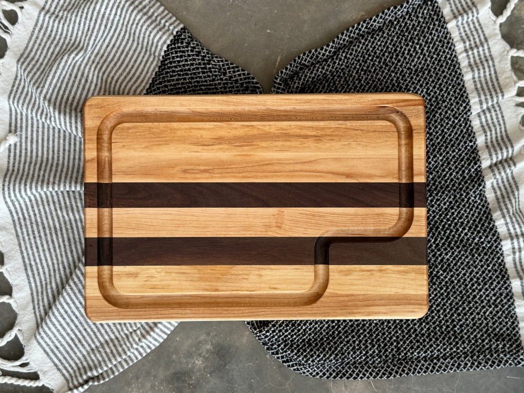 A handcrafted cutting board made from alternating strips of maple and walnut wood, featuring a juice groove for functionality and a smooth, polished finish.