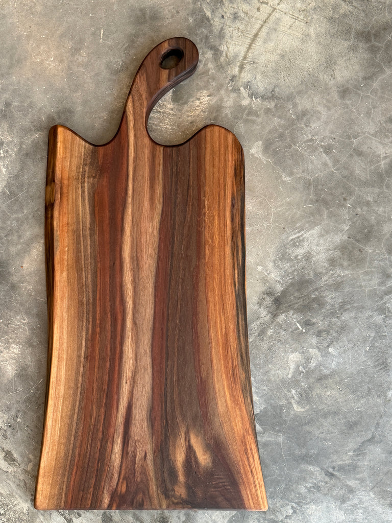 A handcrafted, solid walnut charcuterie board featuring rich natural wood grain, a curved handle with a rounded cutout, and smooth edges, resting on a textured conc