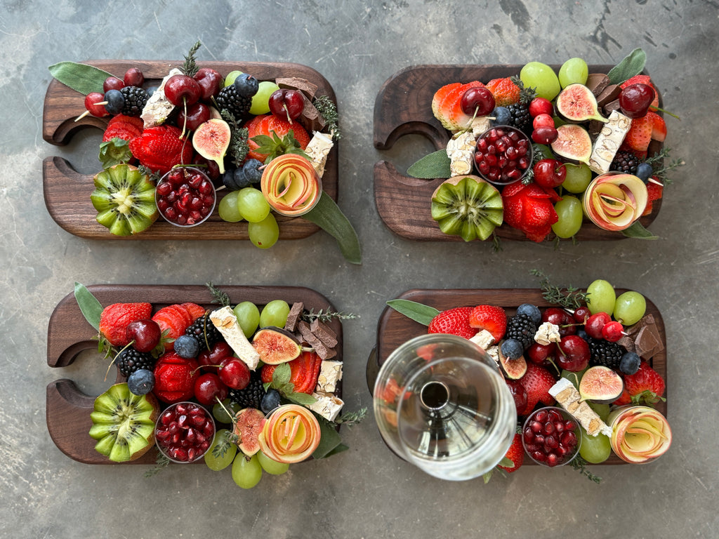 Four wooden black walnut wine and appetizer trays, beautifully arranged with fresh fruits, including strawberries, grapes, figs, cherries, and kiwi, with one tray featuring a wine glass in its designated holder