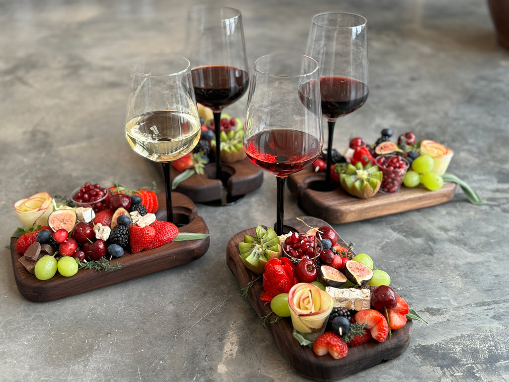 Four individual dark walnut wood appetizer boards with built-in wine glass holders, beautifully arranged with assorted fruits, cheeses, and charcuterie, accompanied by glasses of red and white wine, showcasing an elegant and functional design for gatherings.