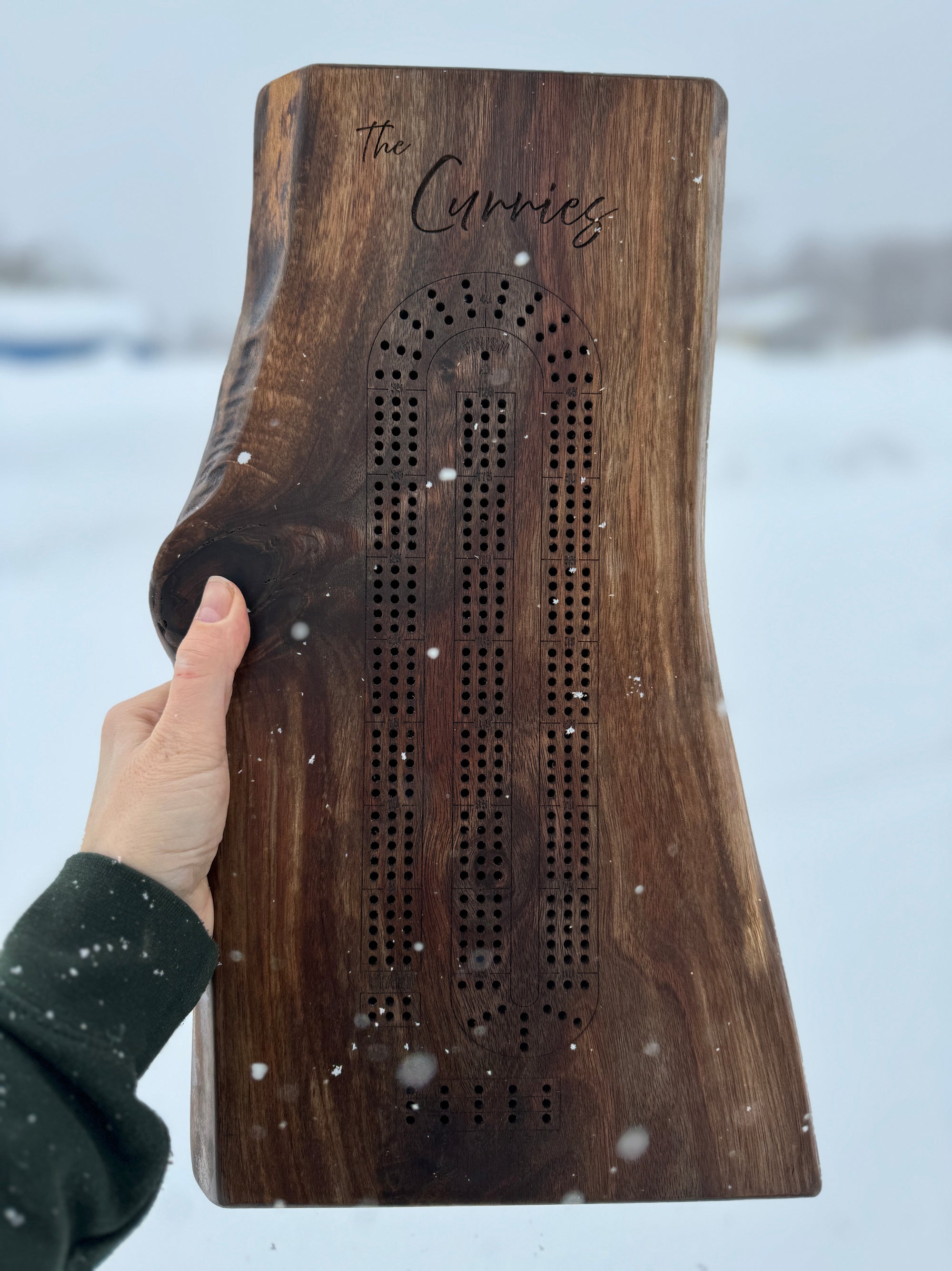 Hand holding a personalized live edge dark walnut cribbage board with 'The Curries' engraving, against a snowy outdoor background, highlighting the craftsmanship and natural beauty of the wood