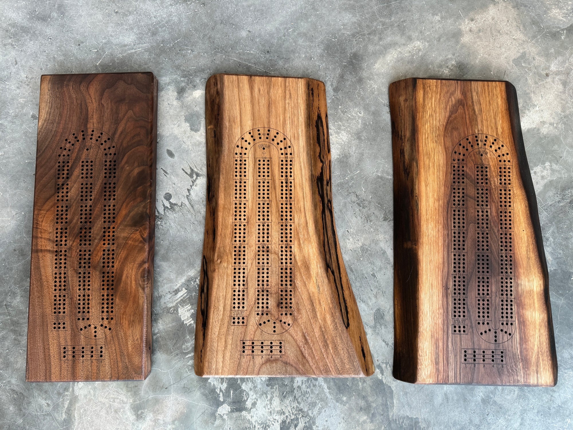 Three handcrafted wooden cribbage boards made from rich walnut, each featuring a unique live edge design. The intricate peg holes and natural wood grain patterns make each board one-of-a-kind. Displayed on a textured concrete surface.