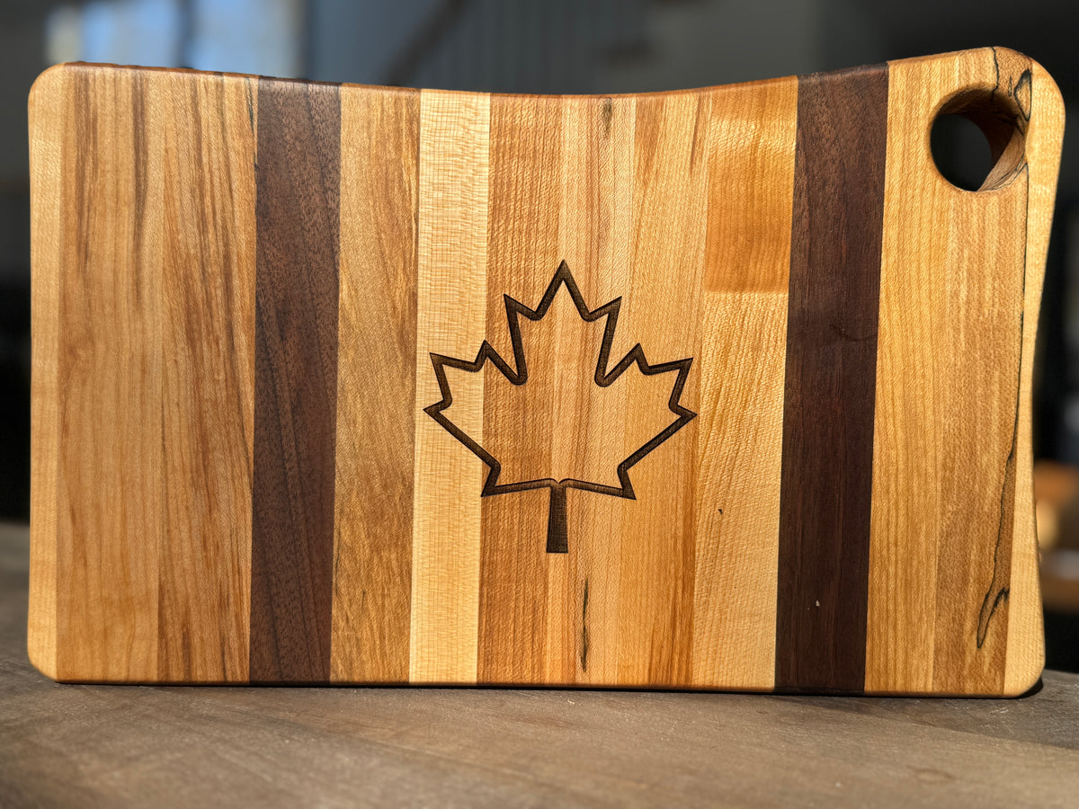 The canadian flag striped maple and walnut cutting board standing on its side with a laser-engraved, outline of maple leaf in the center. The board is sitting on a counter top.