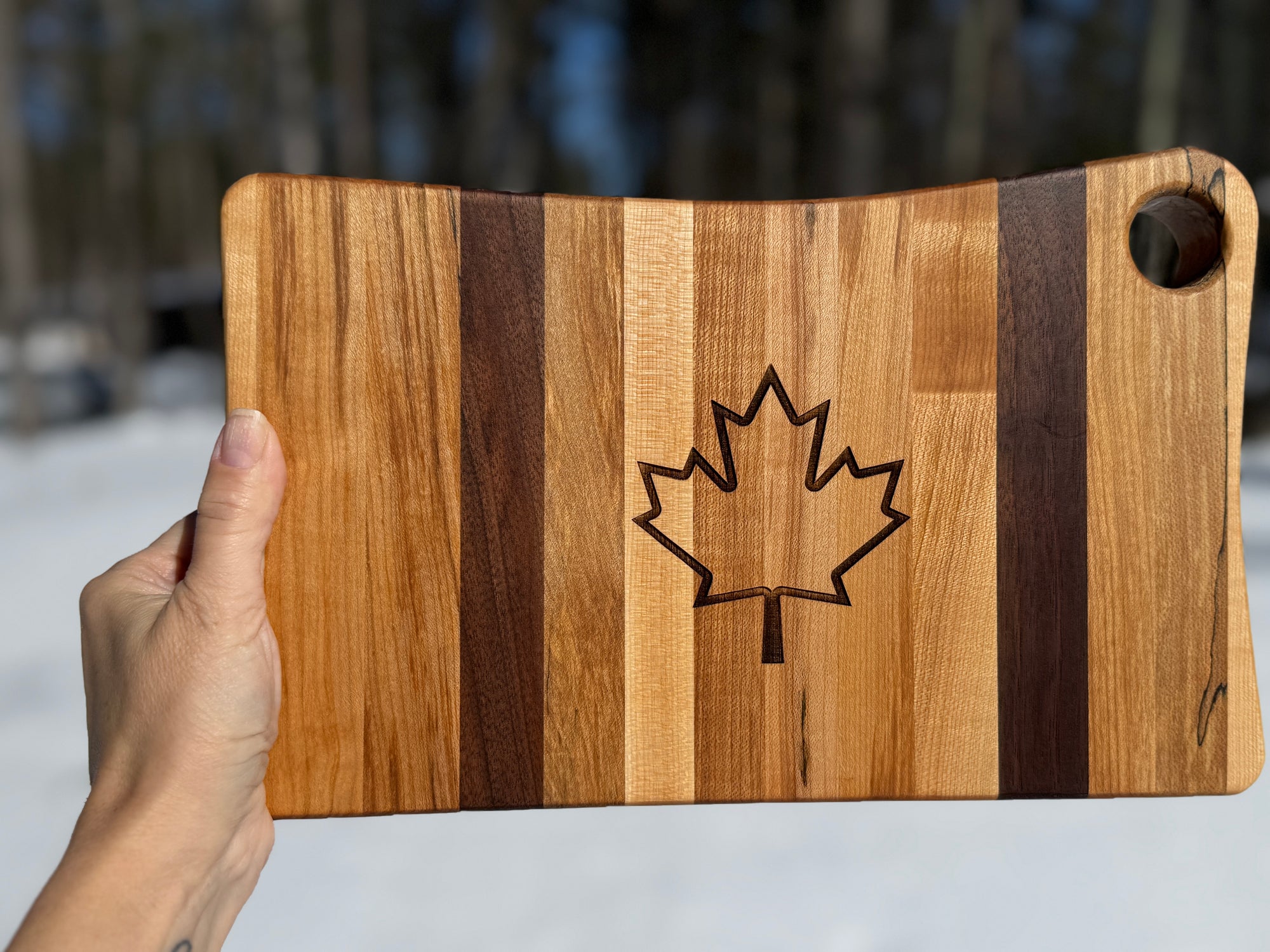 A hand holds up the canadian flag striped maple and walnut cutting board with a laser-engraved, outline of maple leaf in the center, set against a snowy forest background.