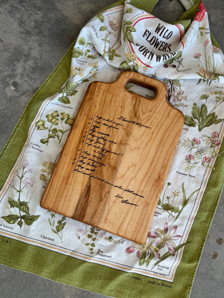 A handwritten brownie recipe is engraved onto a maple cutting board, displayed on a  vintage tea towel.
