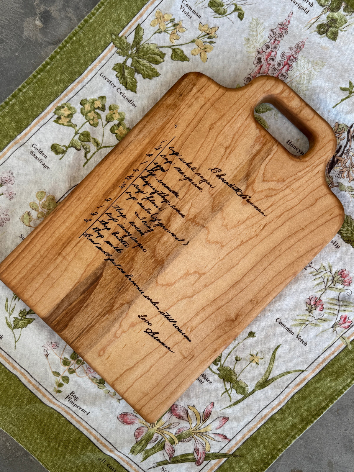 A handwritten brownie recipe is engraved onto a maple cutting board, displayed on a  vintage tea towel. Great personalization gift