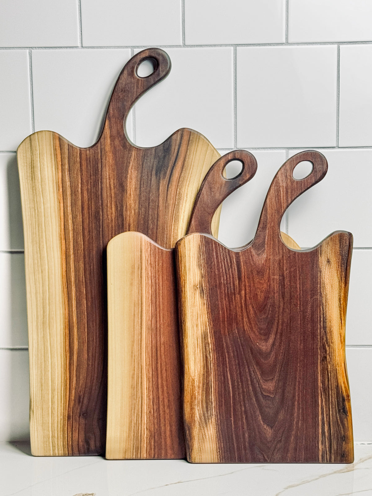 A selection of 3 live edge Black walnut charcuterie boards in varying heights and widths against a white tiled background.