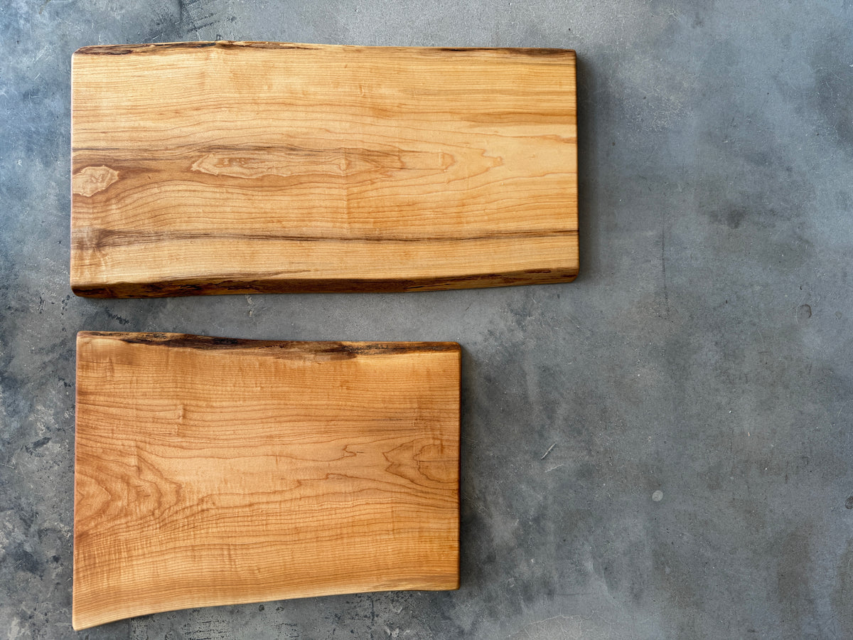 Two wooden maple live edge charcuterie boards lay side by side comparing the two available lengths and showcasing the beautiful wood grain of these large serving platters.