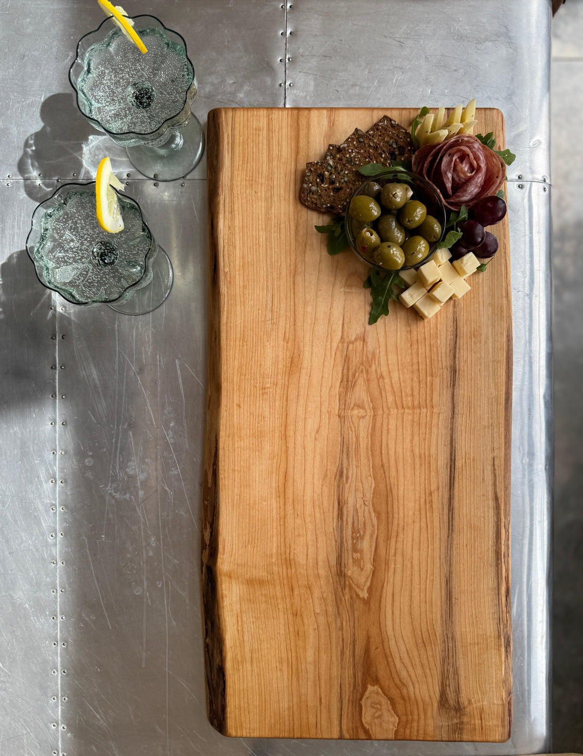 A natural live edge maple charcuterie board. The board is styled with a small arrangement of olives, crackers, cheese cubes, grapes, and salami at the top.