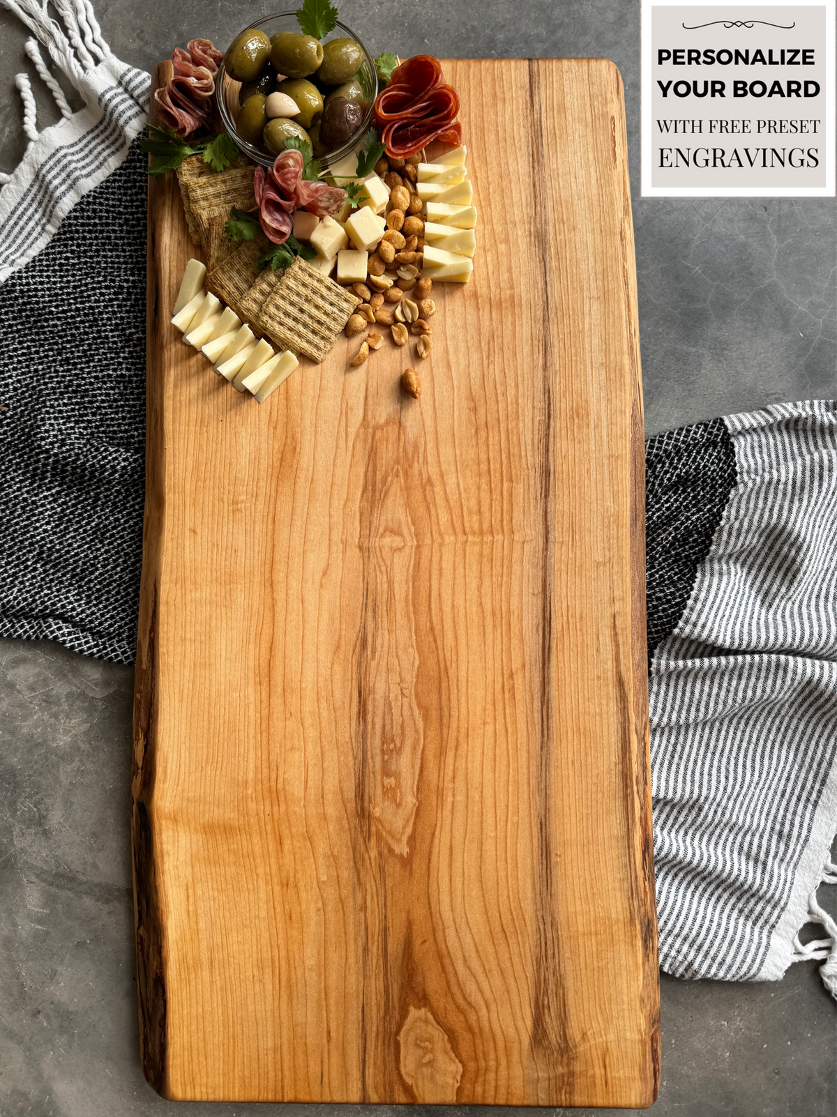 A maple charcuterie board with a natural live edge. The board is styled with a small arrangement of olives, crackers, cheese cubes, grapes, and salami at the top. A black and white striped cloth is partially visible underneath, with a 'Personalize Your Board' engraving promotion displayed in the corner."