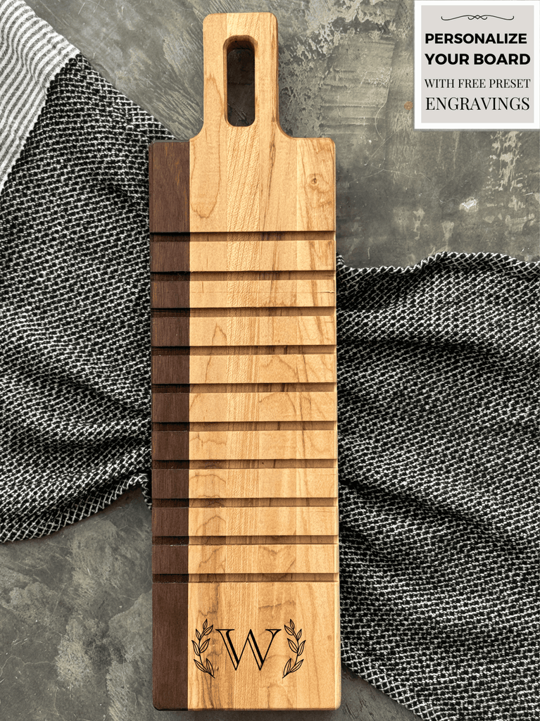 A handcrafted breadboard made with maple and walnut, featuring horizontal grooves and a personalized engraving of the letter W, framed by laurel leaves. The board is displayed on a textured gray surface with striped and checkered cloth accents.