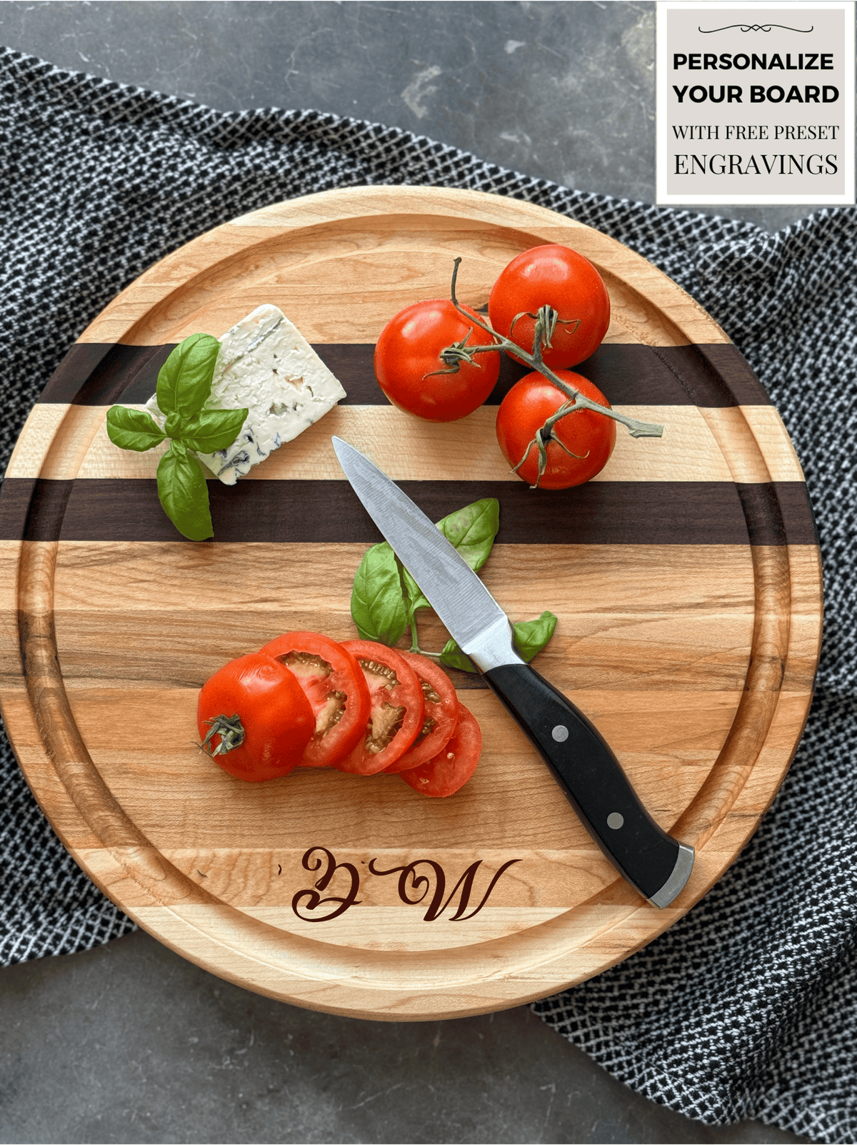 A round, personalized wooden cutting board with maple and walnut stripes, engraved with Intials DW. Fresh tomatoes on the vine, sliced tomatoes, basil, and blue cheese rest on the board. A paring knife lies beside the slices on a textured cloth.
