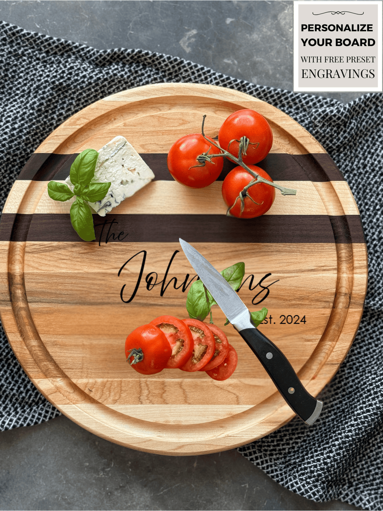 A round, personalized wooden cutting board with maple and walnut stripes, engraved with "The Johnsons Est. 2024." Fresh tomatoes on the vine, sliced tomatoes, basil, and blue cheese rest on the board. A paring knife lies beside the slices on a textured cloth.