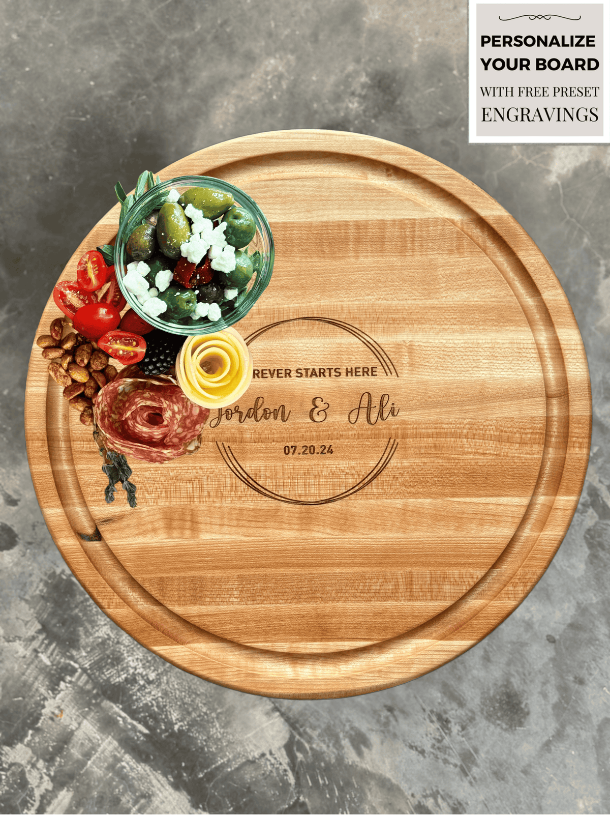 A round, personalized maple wood cutting or charcuterie board engraved with Forever Starts Here - Jordon &amp; Ali - 07.20.24. The board has a juice groove and is topped with olives, feta, cherry tomatoes, salami, a cheese rose, almonds, blackberries, and fresh herbs.