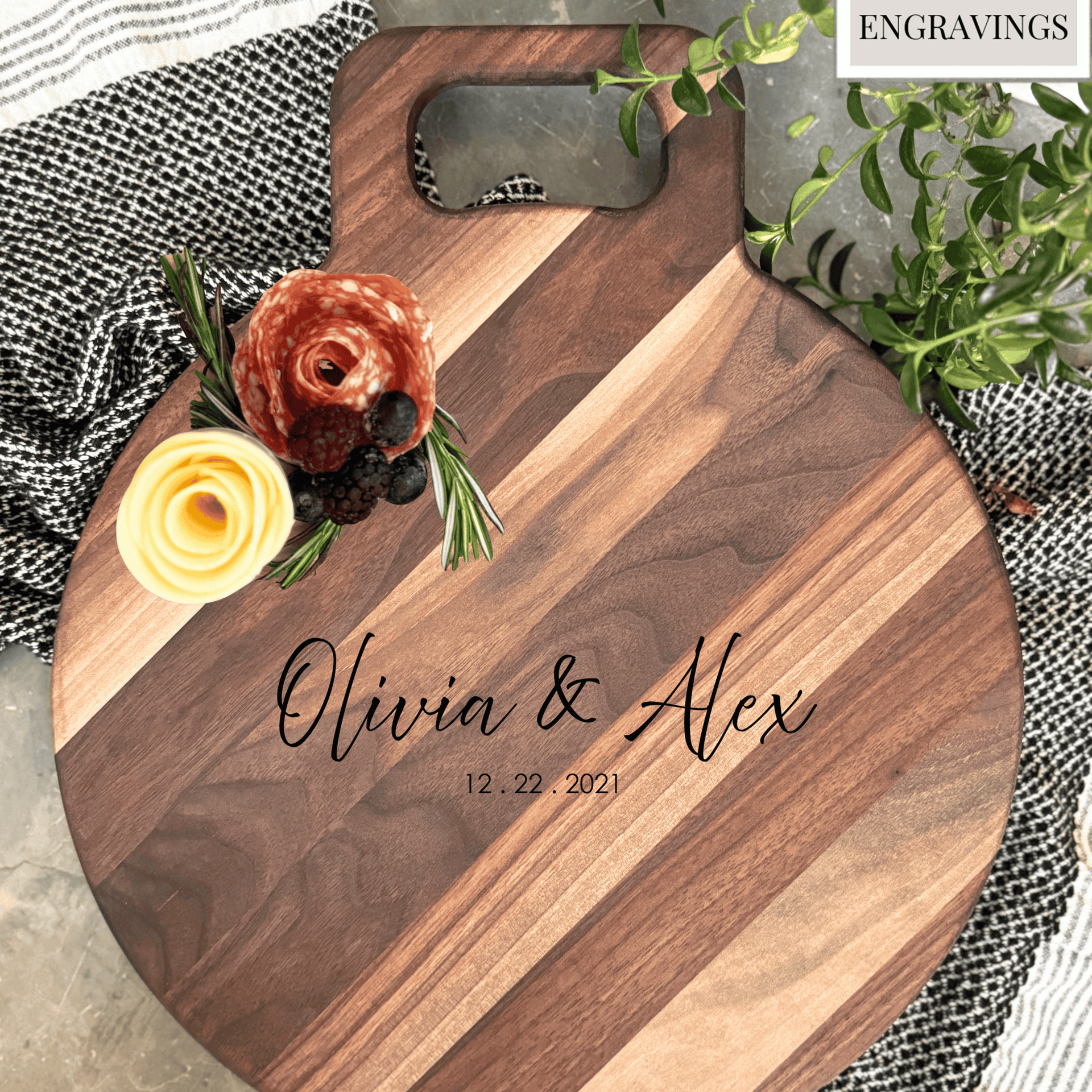 Round wooden charcuterie board with a smooth walnut finish featuring customizable engraving for Olivia & Alex, 12.22.2021. Decorated with neatly arranged salami, cheese, and herbs, perfect for gifting or entertaining. Background includes greenery and textiles.