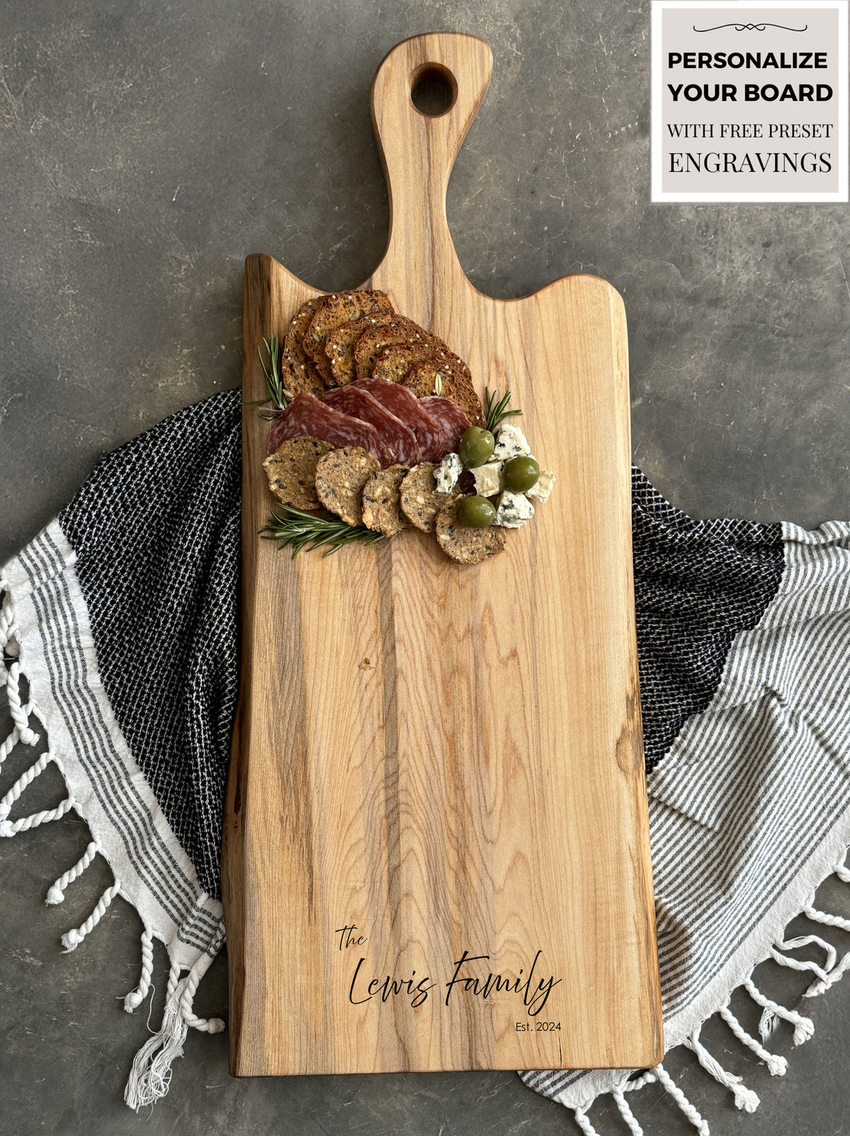 A personalized Maple Live edge charcuterie board engraved with The Lewis Family, Est. 2024, featuring crackers, salami, cheese, olives, and rosemary. The board rests on a striped, fringed towel against a concrete background.