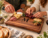 Two people enjoying a charcuterie spread on a handcrafted personalized walnut cutting board. Featuring a custom engraved family monogram, cheeses, cured meats, crackers and fresh fruit. The board includes a juice groove and small rubber feet for stability. A beautiful personalized serving board for gatherings and entertaining.