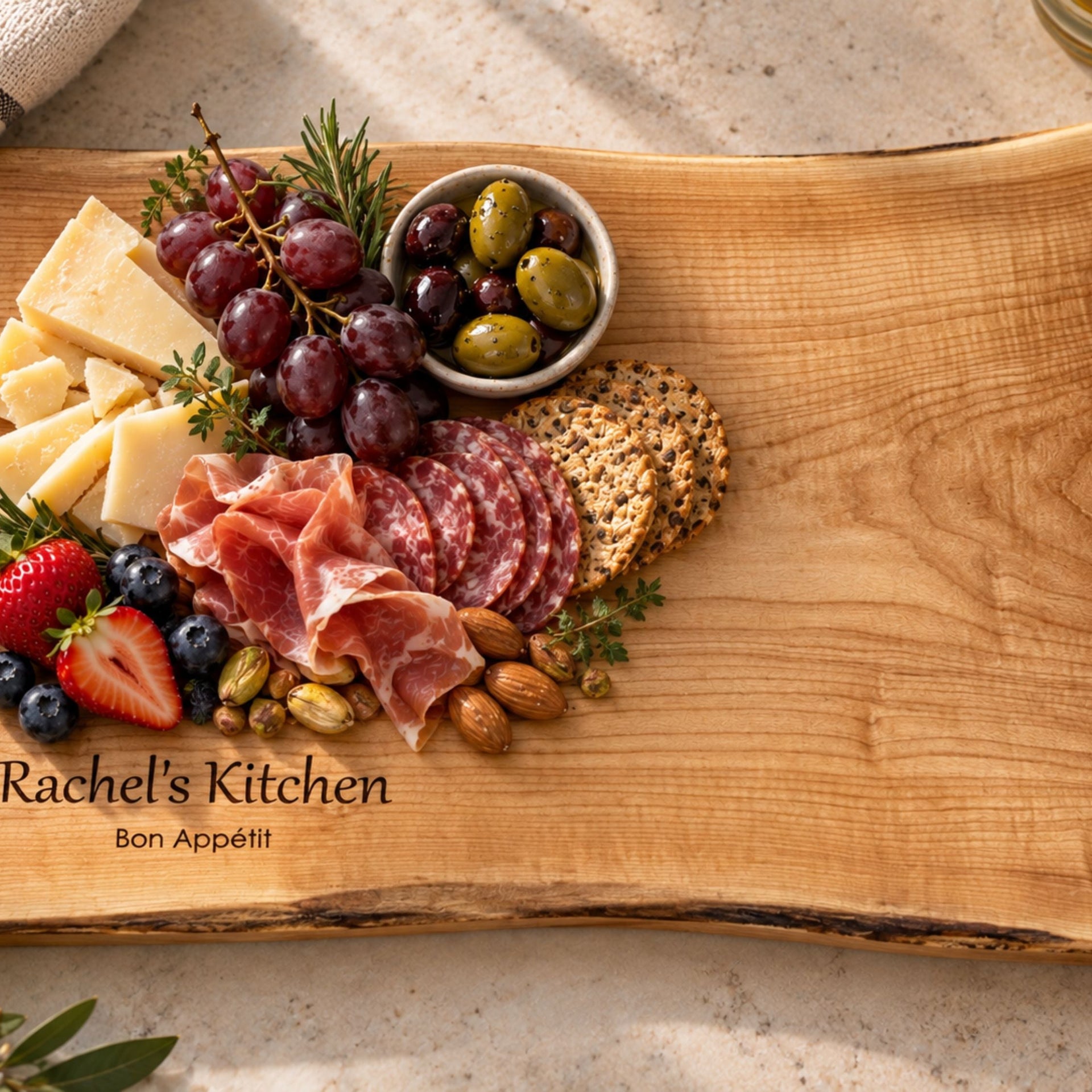 A personalized maple wooden charcuterie board with a live edge, engraved with 'Rachel's Kitchen' and 'Bon Appétit'. The serving board is styled with food items including prosciutto, salami, crackers, grapes, cheese, olives, nuts, and berries on a neutral stone surface.