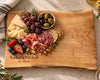A personalized maple wooden charcuterie board with a live edge, engraved with 'Rachel's Kitchen' and 'Bon Appétit'. The serving board is styled with food items including prosciutto, salami, crackers, grapes, cheese, olives, nuts, and berries on a neutral stone surface.