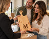 A wooden personalized cutting board with a handle and engraved 'M MILLER EST. 2024' is held up by a woman in a cream sweater while a man in an olive green shirt looks on, with another person in a black top holding the box on the left.