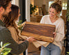 A smiling woman in a cream sweater and jeans holds a large rectangular wooden cutting board Personalized with her last name UNDERWOOD and address engraved below. The charcuterie board is being gifted as a housewarming gift.