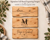 Three personalized wooden board engraving samples are arranged vertically on a light neutral background with decorative greenery and a striped cloth. The boards show sample engraved designs: 'love' with names and date, a monogram 'M' with 'MILLER', and 'Paige & Gabriel' with a heart and date.
