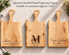 Three personalized wooden cutting boards with handles are arranged on a light surface with decorative greenery and flowers. These are engraving options for gifting to couples for weddings, anniversaries, housewarmings. Text at the top reads, 'Signature Handled Board Engraving Designs Available as a premium upgrade (+$25)'.