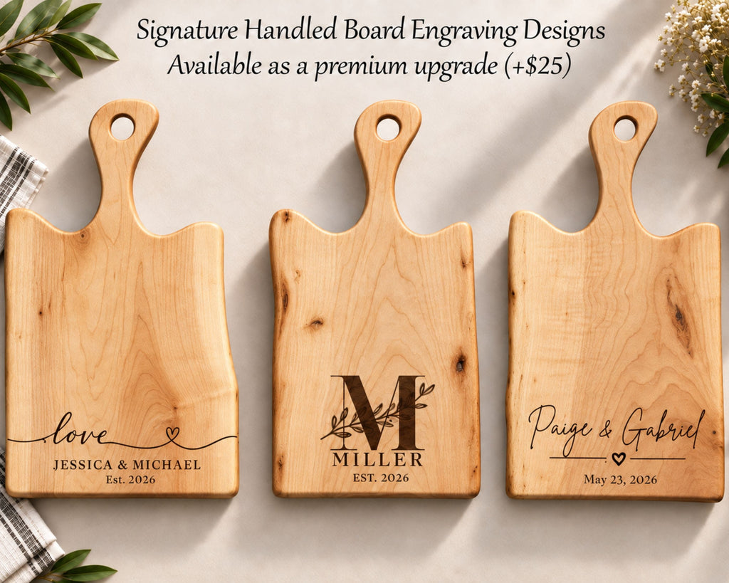 Three personalized wooden cutting boards with handles are arranged on a light surface with decorative greenery and flowers. These are engraving options for gifting to couples for weddings, anniversaries, housewarmings. Text at the top reads, 'Signature Handled Board Engraving Designs Available as a premium upgrade (+$25)'.