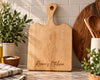 A light natural maple wood cutting board with a handle and the engraved text 'Renee's Kitchen' on the front, displayed on a kitchen counter with plants, a striped cloth, and wooden utensils in the background.