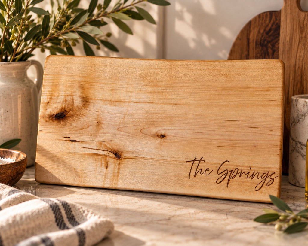 A rectangular natural wood cutting board with the words 'The Springs' engraved in script at the bottom right, styled on a kitchen counter with olive branches, a ceramic pitcher, and a striped cloth.