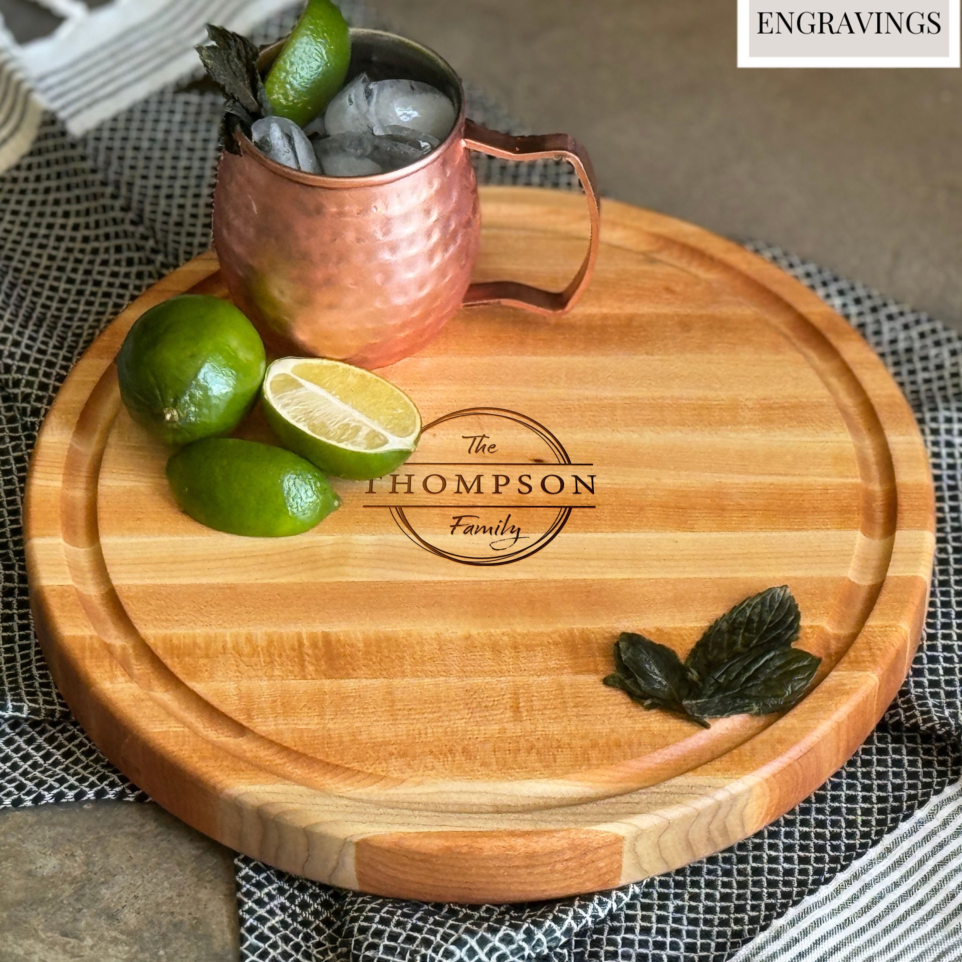 A round, personalized maple wood charcuterie board engraved withThe Thompson Family. The board has a juice groove and is topped with Limes, mint and copper mug of Moscow Mule
